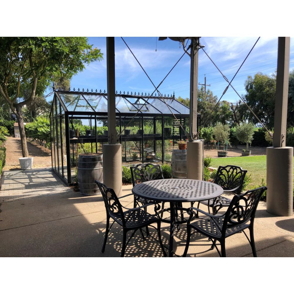 Outdoor patio setting with a round metal table and chairs in front of a greenhouse.