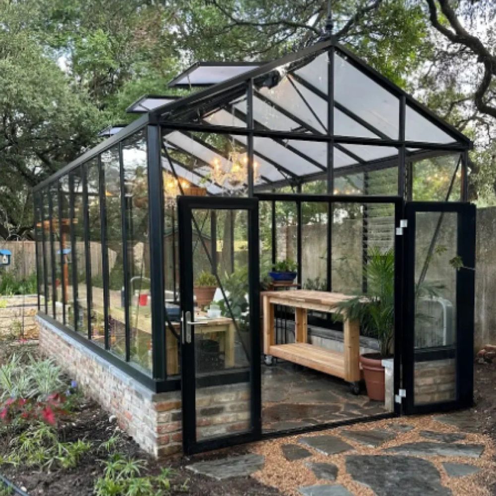 Glass greenhouse with a stone foundation and wooden interior, surrounded by trees and plants.