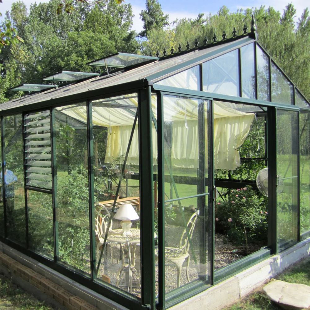 Greenhouse with glass panels and metal frame, surrounded by trees and garden furniture.
